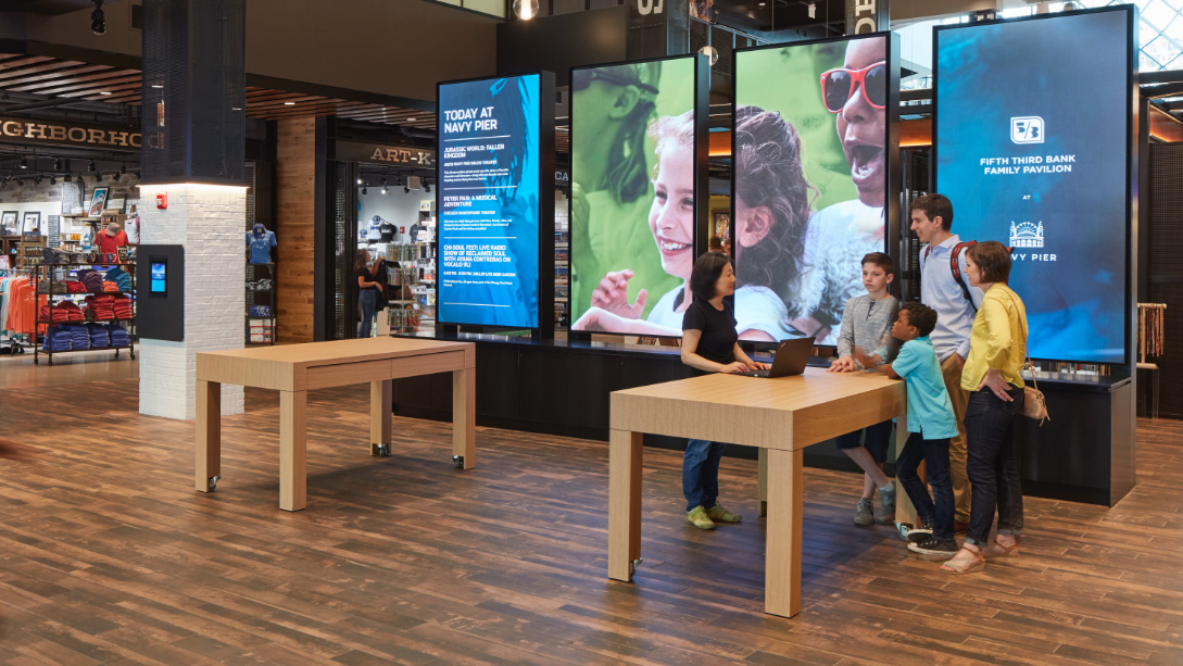 A group of people standing around a table with a person sitting at it.