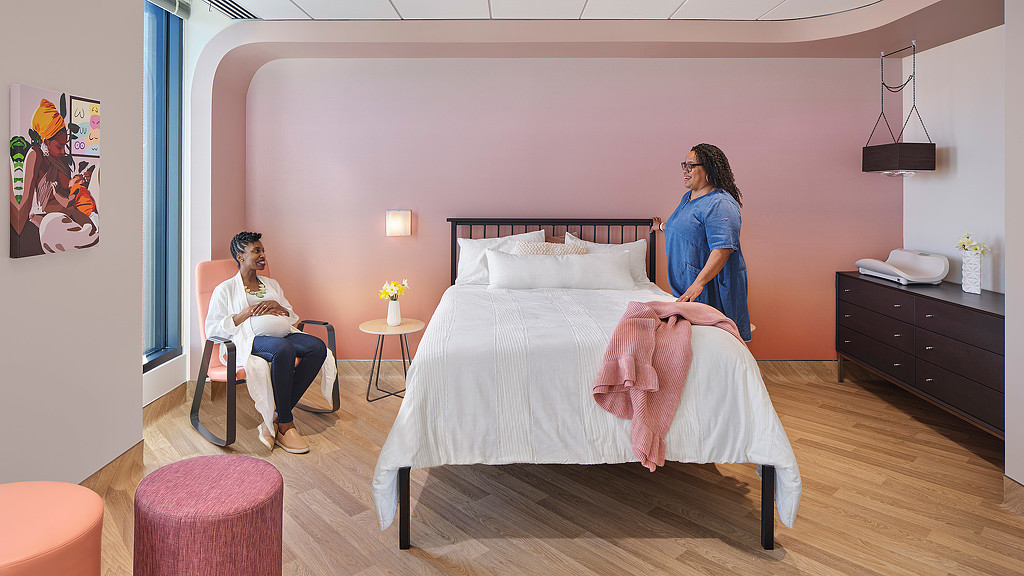 A person standing next to a bed near a pregnant person sitting on a chair.